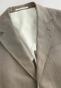 Blazer a righe marrone chiaro con una morbida fodera beige. Presenta rever a punta e una tasca sul petto. Il materiale appare testurizzato e resistente.