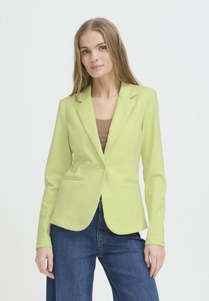 Ung kvinde iført en lys grøn blazer over en brun top og blå jeans, stående foran en ensfarvet hvid baggrund.
