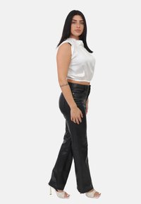 Donna con lunghi capelli scuri che indossa un top senza maniche in satin bianco, pantaloni in pelle nera, tacchi bianchi aperti e un tatuaggio sul suo avambraccio sinistro.