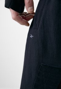 Schwarzer, strukturierter Stoff eines Kleidungsstücks zeigt eine Hand, die in eine Seitentasche greift, mit einem subtilen Logodetail in Grau.