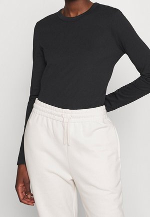 Persona que lleva una camiseta negra ajustada de manga larga y pantalones elásticos de cintura alta en color blanco roto con cordón, contra un fondo liso y claro.