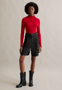 Rotes geripptes Langarmoberteil mit einem rüschengeschnittenen Ausschnitt, kombiniert mit schwarzen knielangen Shorts und schwarzen Absatzstiefeln. Einfaches, maßgeschneidertes Design.