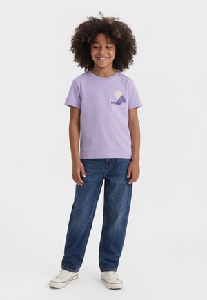 Enfant aux cheveux bouclés souriant, portant un t-shirt lavande avec un logo géométrique, un jean bleu et des baskets blanches, debout devant un fond blanc.