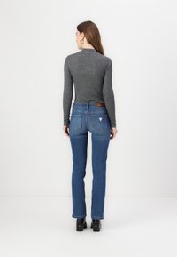 Cămașă lungă de culoare gri închis, purtată cu blugi bleu din denim, având un logo subtil în formă de triunghi pe buzunarul din spate, asortată cu botine negre cu toc.