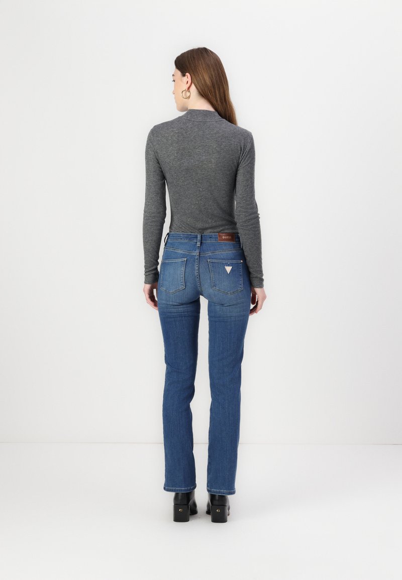 Dunkelgraues Langarmshirt, kombiniert mit blauen Jeans, die ein dezentes Dreieckslogo auf der Gesäßtasche haben, getragen mit schwarzen High-Heels-Stiefeln.