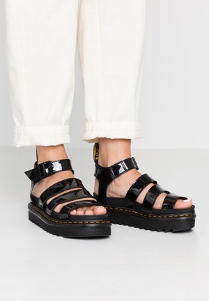 Pieds portant des sandales compensées en cuir verni noir avec plusieurs brides et un pantalon blanc raccourci au-dessus de la cheville.