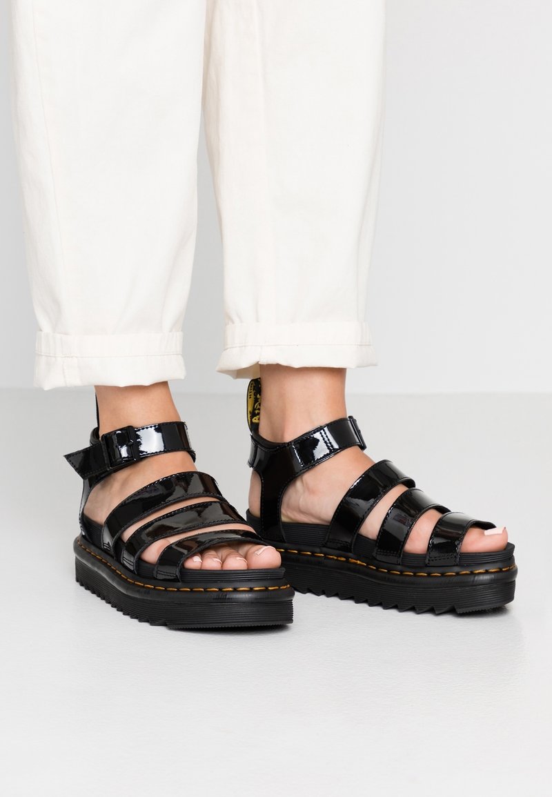 Pieds portant des sandales compensées en cuir verni noir avec plusieurs brides et un pantalon blanc raccourci au-dessus de la cheville.