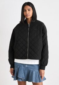Svart quilted bomberjakke med glidelås foran, ribbestrikkede mansjetter og et logo-merke. Brukt over en hvit skjorte og et denimskjørt.