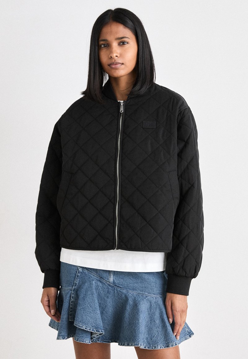 Svart quilted bomberjakke med glidelås foran, ribbestrikkede mansjetter og et logo-merke. Brukt over en hvit skjorte og et denimskjørt.