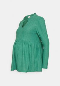 Top de maternidade verde, de mangas longas, com decote em V e colarinho com babados. Apresenta um tecido texturizado e um ajuste relaxado e evasé na barriga.