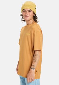 Kortærmet, sennepsfarvet t-shirt lavet af blød bomuld, med et diskret logo på brystet. Båret med en gul strikket hue.