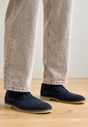 Zapatos de ante azul marino oscuro con punta redondeada, costuras mínimas y suela de goma clara, combinados con jeans beige claros sobre un suelo de madera.