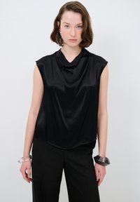 Top senza maniche in satin nero con scollatura drappeggiata, texture liscia, abbinato a pantaloni neri e bracciali trasparenti.