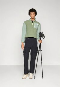 Zeleni pulover iz flisa s polovičnim zadrgo in žepom na prsih, v kombinaciji z mornarsko modrimi cargo hlačami in lahkimi bež pohodniškimi čevlji. V roki drži pohodniške palice.