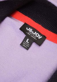 Sweatertag met zwarte stof en witte tekst "J&JOY," maat "L," en een rode streep tegen een lavendel achtergrond.
