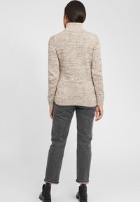 Strikkegenser i beige med høy krage, med et strukturert mønster. Kombinert med grå rette bukser og ankelstøvler.