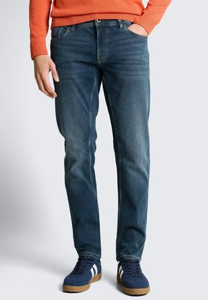 Mann trägt verwaschene blaue Jeans und marineblaue Sneakers mit weißen Streifen, orangener Pullover auf Taillenhöhe sichtbar, steht vor einfachem Hintergrund.