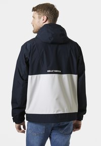 Marineblauwe en witte kleurblokkering waterdichte jas met capuchon, voorzien van elastische manchetten en het "HELLE HANSEN" logo op de achterkant.