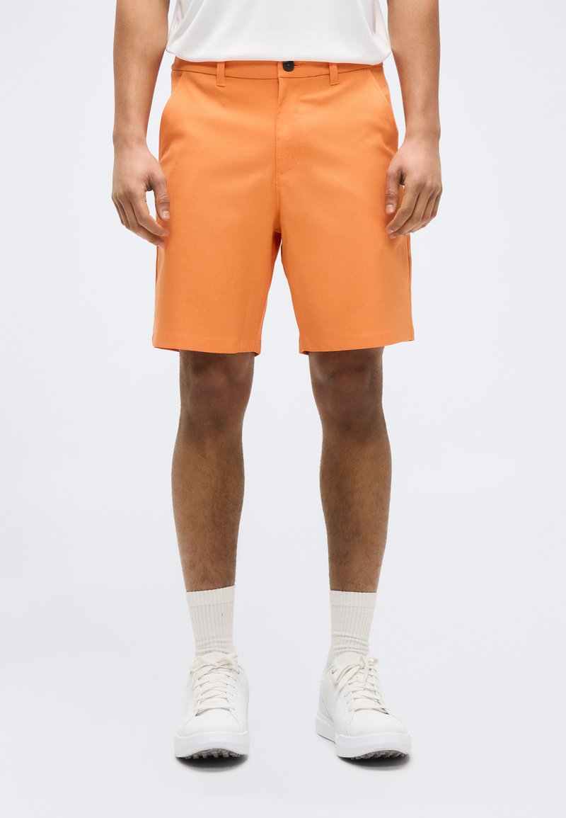 Άνδρας που φοράει φωτεινά πορτοκαλί βερμούδες μέχρι το γόνατο, λευκές σκουρόχρωμες κάλτσες και άσπρα αθλητικά παπούτσια, στέκεται μπροστά σε απλό φωτεινό φόντο.