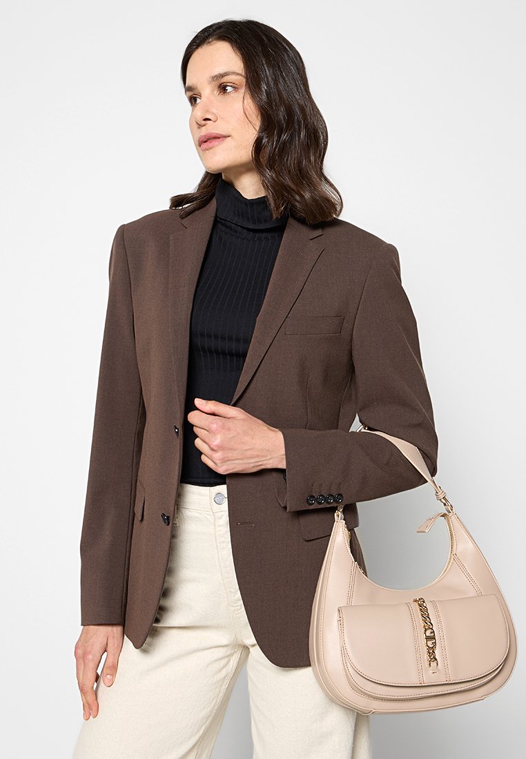 Donna con capelli castani lunghi fino alle spalle, che indossa un blazer marrone su un dolcevita nero, mentre tiene una borsa beige e pantaloni bianco sporco.