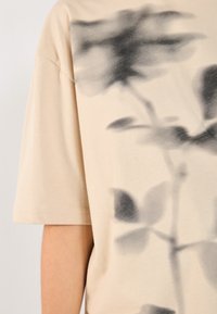 Camiseta de manga corta beige con estampado gráfico de flores en negro. Tejido de algodón suave con un corte relajado. Detalles de costura en el dobladillo y las mangas.
