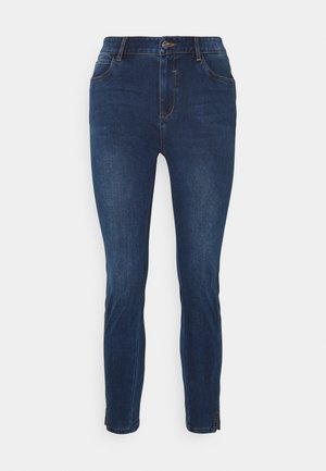 Jeans slim-fit bleu foncé avec poches avant, fermeture à bouton et zips aux chevilles sur un fond blanc.
