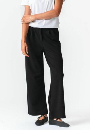 Žena stojaca s prekriženými rukami, oblečená v bielej krátkorukávovej košeli, čiernych širokých nohaviciach a čiernych topánkach štýlu Mary Jane.