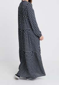 Kobieta w długiej, czarnej sukni z długim rękawem i długości do kostek, w białe kropki, w czarno-białych sneakersach, stojąca bokiem na jednolitym tle.