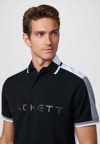 Camisa polo negra con un gráfico gris del logo "HACKETT". Presenta paneles de hombros blancos en contraste y un cuello clásico. Textura de tela suave.