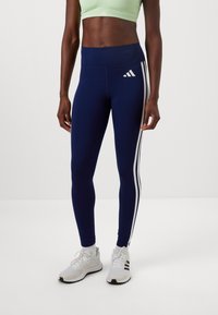 Marineblauwe leggings van rekbare stof met een hoge tailleband, met witte zijstrepen en een Adidas-logo. Gecombineerd met witte sneakers.