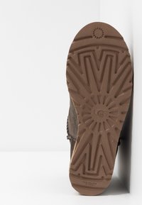 Suela de bota marrón con un profundo patrón de zigzag apoyada contra una pared blanca, mostrando un agarre texturizado y el logo de la marca.