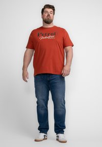 Camiseta de algodón roja con gráfico de "Petrol Industries", combinada con jeans azules y zapatillas blancas con acentos negros. El modelo está de frente.
