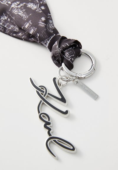 Porte-clés avec une étiquette "Karl" noire et argentée attachée à une écharpe noire nouée avec un design imprimé blanc, et un anneau en argent.