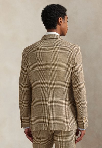 Uomo visto di spalle che indossa una giacca da completo a quadri beige e marrone con pantaloni abbinati, su uno sfondo neutro e semplice.