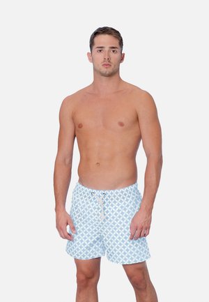 Junger Mann steht, oberkörperfrei, trägt hellblaue und weiße Badeshorts mit Muster und Kordelzug vor einfachem Hintergrund.