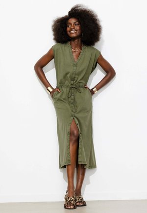 Femme avec coiffure afro sourit, portant une robe sans manches vert olive boutonnée avec une fente avant et des sandales à imprimé léopard, mains dans les poches.