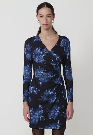 Mujer de pie vistiendo un vestido envolvente negro de manga larga con un patrón floral azul y pendientes de aro plateados, contra un fondo liso.