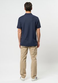 Bărbat stând cu fața spre spate, purtând un tricou polo bleumarin, pantaloni bej și adidași albi, pe un fundal simplu deschis.