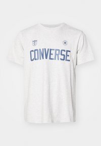 Hallikas puuvillane t-särk, millel on sinine "CONVERSE" tekst, logo plekk vasakul rinnal ja ümar embleem paremal. Lühikesed varrukad, ümar kaelus.