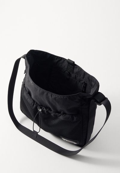 Sac en nylon noir avec une poche en maille texturée, une bandoulière réglable et un cordon de serrage. L'intérieur est doublé avec un tissu lisse.