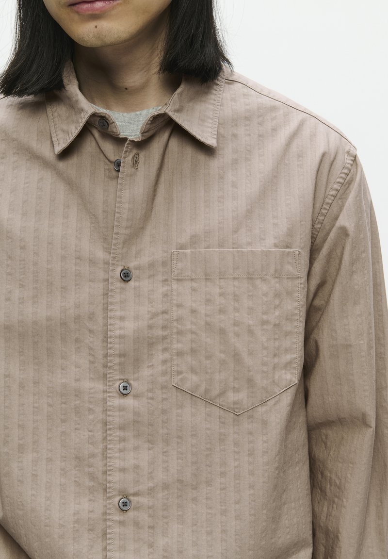 Chemise rayée beige avec une fermeture à boutons, une poche poitrine, et une texture douce. Comprend des boutons gris et un col.