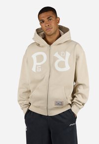 Beige zip-up hette med store hvite logobokstaver foran, kengurulomme og ribbestrikkede mansjetter. Laget av myk stoff med en avslappet passform.