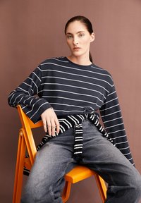 Camicia a maniche lunghe blu navy con righe bianche, indossata sopra jeans scuri e una cravatta a righe bianco e nere. Modello seduto su una sedia arancione.