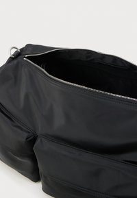 Fekete nejlon keresztpántos táska két első zsebbel és félig nyitott ezüst cipzárral, amely a üres belsejét mutatja.