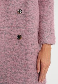 Gros plan sur un manteau en laine texturée rose avec de grands boutons foncés et la main droite d'une personne reposant le long de la manche.