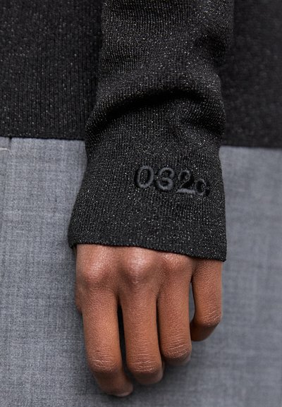 Must musta, sädelev must pullover, millel on sooniline tekstuur ja tikand "032c" detailiga varrukal, kantud halli kangas taustal.