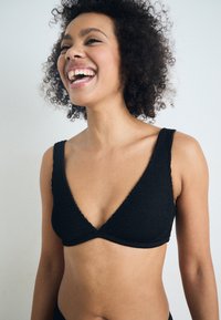 Czarny, dziany biustonosz typu bralette z głębokim dekoltem w kształcie litery V, szerokimi teksturowanymi ramiączkami i bezszwowymi brzegami, zaprojektowany z myślą o komforcie i elastyczności.