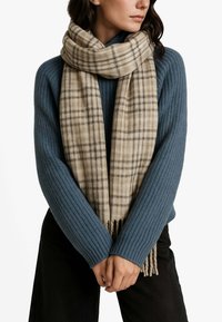Sciarpa a quadri beige con estremità frangiate, realizzata in tessuto morbido, avvolta attorno a un maglione blu a coste. Il design presenta linee marroni scure.