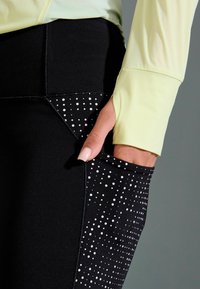 Des leggings noirs présentent un design texturé avec de petits points blancs sur un côté et une poche élégante. Associés à un haut à manches longues jaune pâle.