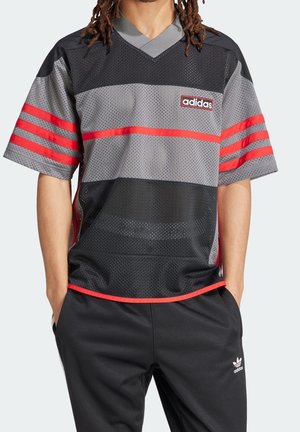 Persoon die een zwart-grijs Adidas mesh shirt met rode strepen en zwarte Adidas broek draagt, handen in de zakken.